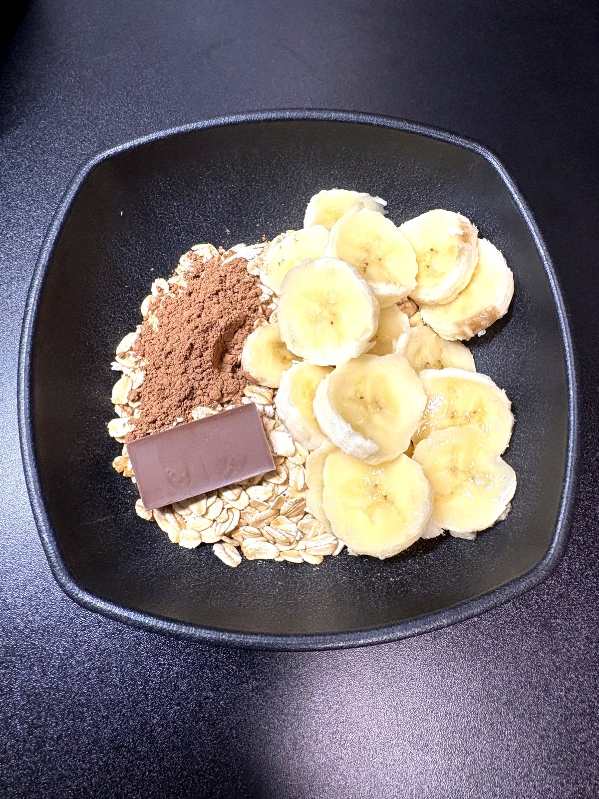 オートミール毎日レシピの材料。ロールドオーツ・バナナ・純ココア・ミルクチョコを黒い皿に並べた状態